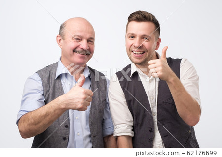 Two caucasian mature men father and son holding thumbs up approving your choice. Two caucasian mature men father and son holding thumbs up approving your choice. 62176699