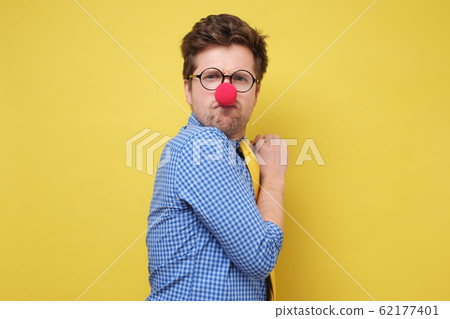 A young greedy man with red clown nose hides something in his hands and looks warily forward A young greedy man with red clown nose hides something in his hands and looks warily forward 62177401