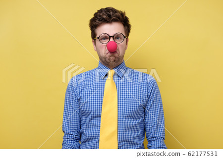 Caucasian young man in red clown nose being irritated and sad. 62177531