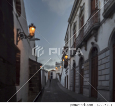 Defocused evening empty narrow street at with glowing lanterns, houses in traditional colonial architecture and cobble stone paving Defocused evening empty narrow street at with glowing lanterns, houses in traditional colonial architecture and cobble stone paving 62203704