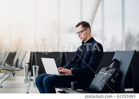 A business man sitting at the airport park working with his laptop and drinking takeaway coffee while waiting the flight departure. A business man sitting at the airport park working with his laptop and drinking takeaway coffee while waiting the flight departure. 62205065