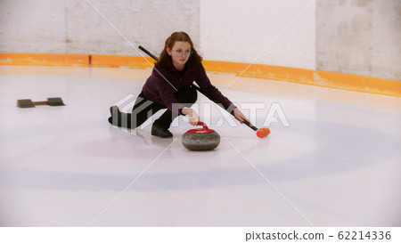 A young woman in glasses with short hair pushes off in the ice field with a granite stone 62214336