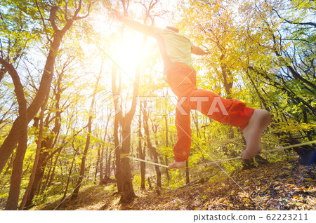Wide angle male tightrope walker balancing barefoot on slackline in autumn forest. The concept of outdoor sports and active life of people aged 62223211