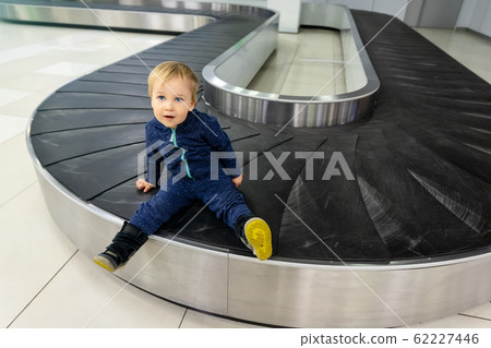 Alone cute caucasian blond toddler boy sitting on empty airport arrival terminal luggage conveyor belt. Lost found child. Travel by plane with children. Safety rules. Delay flight. Waiting baggage Alone cute caucasian blond toddler boy sitting on empty airport arrival terminal luggage conveyor belt. Lost found child. Travel by plane with children. Safety rules. Delay flight. Waiting baggage 62227446