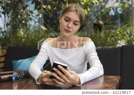 Cheerful blond woman blogger is reading incoming sms message on smartphone connected to free wifi in cafe. Positive young student watching broadcasting online on modern mobile phone. 62229617