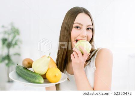 Woman holding fruits assortment served on white plate against light background. Closeup portrait of cheerful young woman with perfect teeth biting green apple. Healthy, beauty concept 62230472