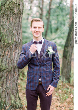 Trendy man groom in stylish suit touching his bow tie posing on the background of pine forest Trendy man groom in stylish suit touching his bow tie posing on the background of pine forest 62240603