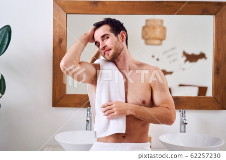 Happy brunet male with towel on shoulder stands near mirror in bath 62257230