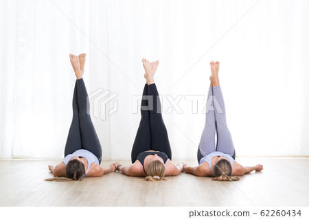 Group of three young sporty attractive women in yoga studio, lying on the floor holdings legs up against white background. Healthy active lifestyle, working out indoors in gym. Group of three young sporty attractive women in yoga studio, lying on the floor holdings legs up against white background. Healthy active lifestyle, working out indoors in gym. 62260434