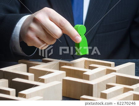 A male businessman holds a green figure over a maze. Make a path to success, provide support. Pass test, complete task. Professional skills development. Find a way out of the situation. Reach the goal 62270246