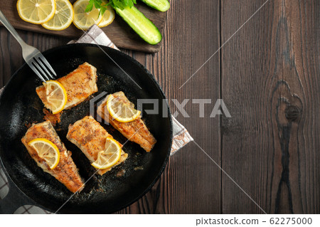 Portioned pieces of fried fish in a black cast-iron pan with fresh cucumbers and herbs. Traditional dinner of a village fisherman, top view, flat lay, copy space Portioned pieces of fried fish in a black cast-iron pan with fresh cucumbers and herbs. Traditional dinner of a village fisherman, top view, flat lay, copy space 62275000