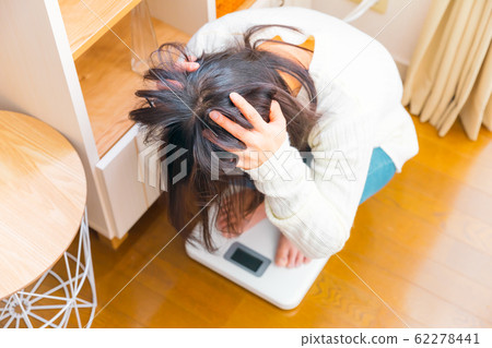A woman riding a weight scale, holding her head 62278441
