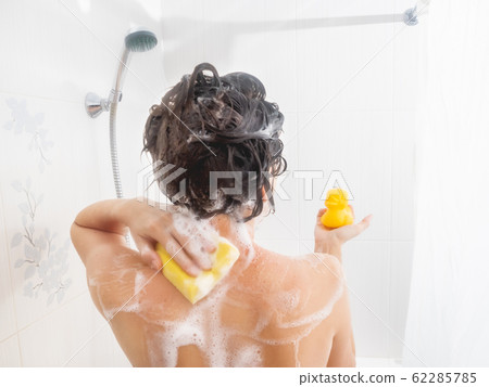 Naked woman with short hair takes a shower with funny yellow rubber duck. Woman washes her shoulders with yellow sponge. White bathroom. Naked woman with short hair takes a shower with funny yellow rubber duck. Woman washes her shoulders with yellow sponge. White bathroom. 62285785