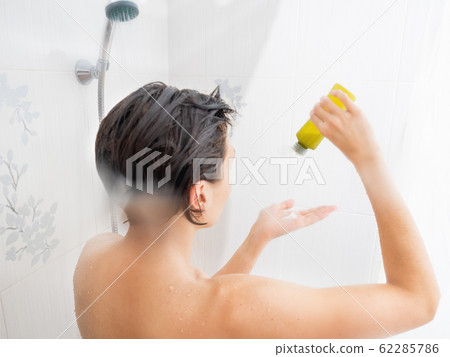 Naked woman with short hair takes a shower. Woman squeezes shampoo out of a green bottle.  White bathroom. 62285786