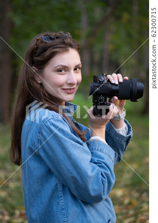 Cute hipster photographer girl takes a picture on a professional camera and smiles. Portrait in the autumn park, against the background of trees and fallen leaves 62300715
