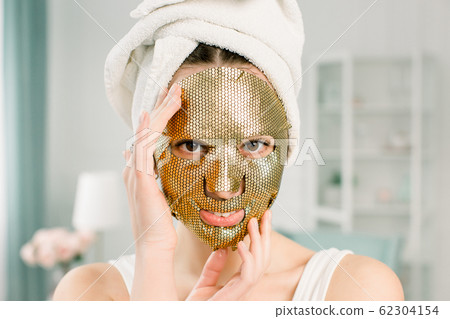 Woman Gold Sheet Mask. Beautiful young woman in white towel on the head with Golden Skin Cosmetic, looking at camera. Beauty Skincare and Treatment Woman Gold Sheet Mask. Beautiful young woman in white towel on the head with Golden Skin Cosmetic, looking at camera. Beauty Skincare and Treatment 62304154