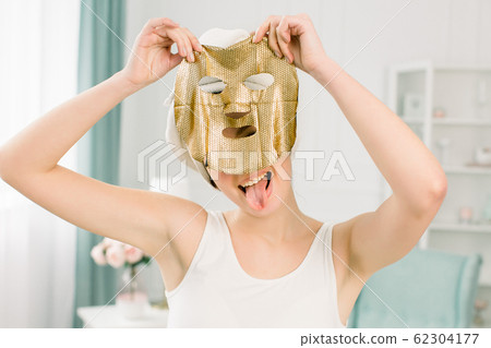 Cosmetic procedure, pretty funny girl in white towel with gold sheet mask on light background. Beauty, spa, skin and body care. 62304177