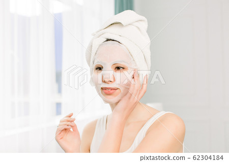 Closeup portrait of young pretty woman applying paper sheet mask on her face. Cosmetic procedure. Beauty spa and cosmetology. Closeup portrait of young pretty woman applying paper sheet mask on her face. Cosmetic procedure. Beauty spa and cosmetology. 62304184