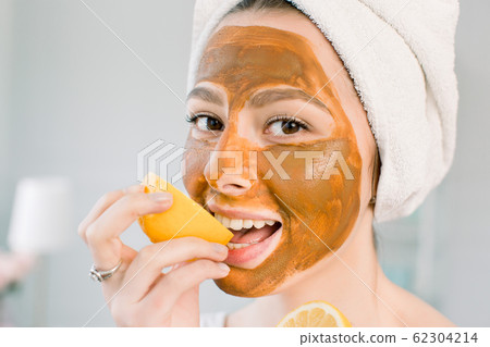 Attractive young woman with white towel on the head and with mud facial brown mask bites a lemon and smiles. Beauty, spa, skin and body care. Attractive young woman with white towel on the head and with mud facial brown mask bites a lemon and smiles. Beauty, spa, skin and body care. 62304214