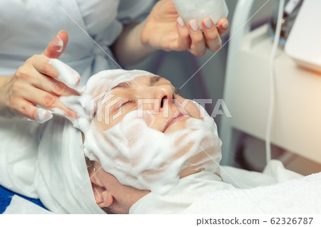 Close-up beautician doctor hands making anti-age procedures, applying foam cleansing mask for mid-aged female client at beauty clinic. Cosmetologist doing skincare treatment .Health care therapy 62326787