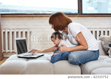 A woman with a child at a laptop sitting on a sofa. Work at home, freelancer, work during maternity leave for remote access. A woman with a child at a laptop sitting on a sofa. Work at home, freelancer, work during maternity leave for remote access. 62335752
