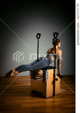 Young girl doing pilates exercises with a reformer bed. Beautiful slim fitness trainer on reformer gray background, low key, art light. Fitness concept Young girl doing pilates exercises with a reformer bed. Beautiful slim fitness trainer on reformer gray background, low key, art light. Fitness concept 62335778