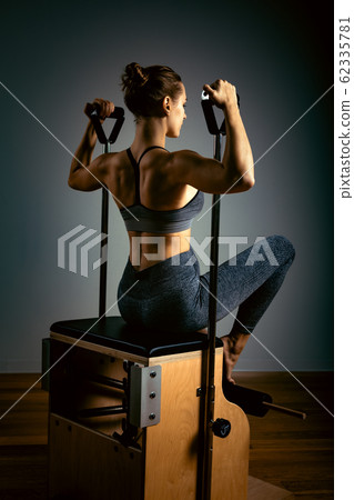 Young girl doing pilates exercises with a reformer bed. Beautiful slim fitness trainer on reformer gray background, low key, art light. Fitness concept 62335781