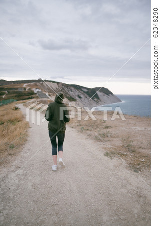 Concentrated slim woman jogging on a cliff above the sea at sunset in cloudy weather. Endurance training, preparation for a marathon Concentrated slim woman jogging on a cliff above the sea at sunset in cloudy weather. Endurance training, preparation for a marathon 62348290