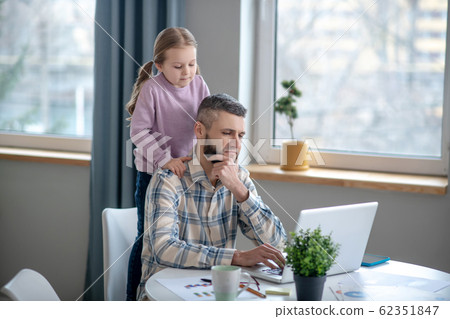 Seated focused dad, daughter standing behind, they are looking into laptop. 62351847