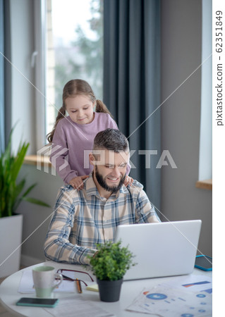 Dad sitting working at laptop, little daughter behind daddys back. 62351849