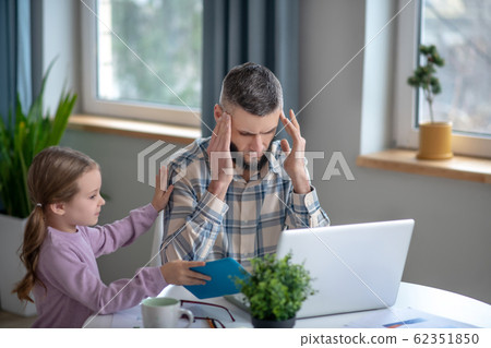 Tired dad sitting at laptop covering his eyes and daughter. 62351850