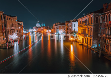 Venice, Italy night scenery of Grand Canal. Vivid light trails of ferries and boats reflected on water surface. Majestic Basilica di Santa Maria della Salute in background Venice, Italy night scenery of Grand Canal. Vivid light trails of ferries and boats reflected on water surface. Majestic Basilica di Santa Maria della Salute in background 62358270