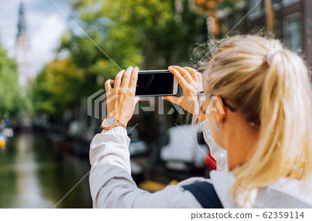 Woman tourist taking a picture of canal in Amsterdam on the mobile phone. Warm gold afternoon sunlight. Travel in Europe 62359114
