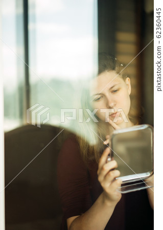 Young woman standing by the window with a mirror in her hands Young woman standing by the window with a mirror in her hands 62360445