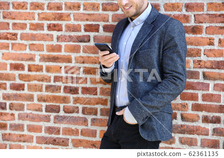 Young casual business man using smartphone in urban city background relaxing on brick wall texting sms on phone app living a modern lifestyle. Closeup of unrecognizable person 62361135