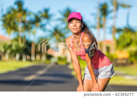 Tired runner woman ready for running wearing sports cap and sport armband with earphones listening to mobile music. Active fit Asian girl resting taking a jogging break on outdoor summer street 62369277