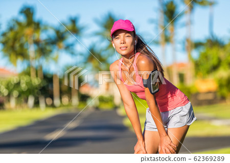 Tired runner woman ready for running wearing sports cap and sport armband with earphones listening to mobile music. Active fit Asian girl resting taking a jogging break on outdoor summer street 62369289