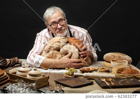 Happy old baker looking at camera and smiling while hugging loaves of bread Happy old baker looking at camera and smiling while hugging loaves of bread 62383277