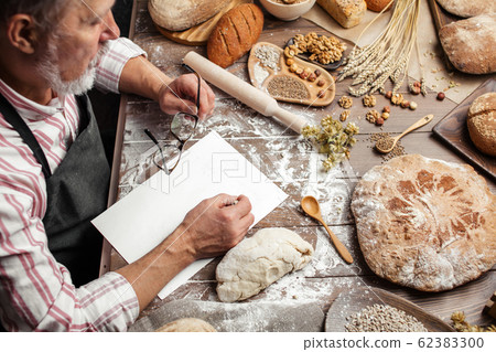 Old Baker writing down old-time recipe in bakery notebook surrounded by bread Old Baker writing down old-time recipe in bakery notebook surrounded by bread 62383300