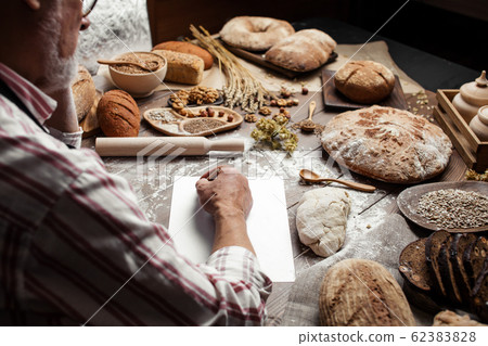 Old Baker writing down old-time recipe in bakery notebook surrounded by bread Old Baker writing down old-time recipe in bakery notebook surrounded by bread 62383828