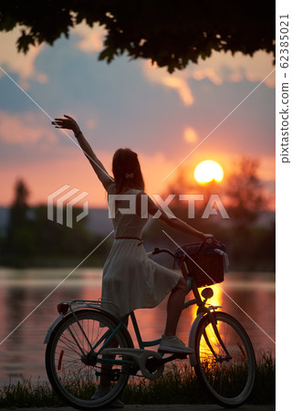 Rear view of girl with retro bike on which the basket enjoys the fiery sun at sunset 62385021