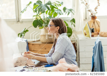 Smiling young dressmaker woman sews clothes on a sewing machine in her workshop 62385080