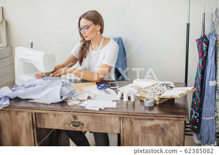 A young dressmaker woman sews clothes on a sewing machine. Smiling seamstress in his workshop 62385082
