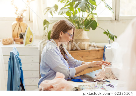 Smiling young dressmaker woman sews clothes on a sewing machine in her workshop Smiling young dressmaker woman sews clothes on a sewing machine in her workshop 62385164