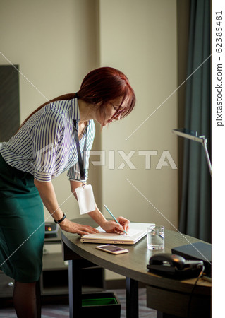 Red-haired female designer noting details of work during phone conversation. 62385481