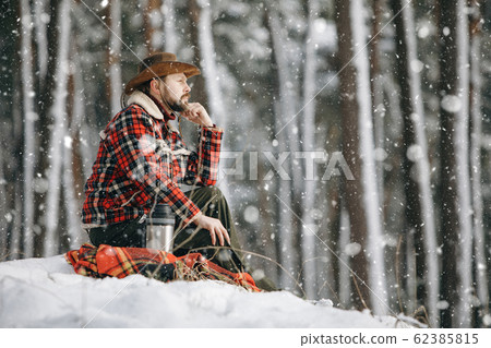Hiker in Checkered Jacket Sitting on a Small Snowy Forest Hill 62385815