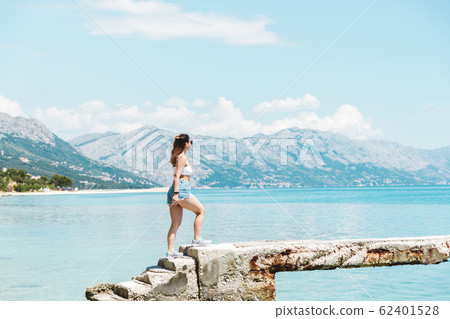 time to travel. beautiful woman in white topic and jeans shorts alone on seaside looking to the blue ocean and enjoying the view 62401528