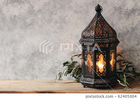 Black ornamental Moroccan, Arabic lantern. Green olive leaves, branches on old wooden table, blurred grunge wall background,. Greeting card for Muslim holiday Ramadan Kareem. Festive still life. 62405804
