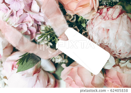 Wedding, birthday stationery mock-up scene. Empty gift tag, label with pink silk ribbon. Decorative floral composition. Closeup of pink roses petals, peonies and hydrangea flowers. Selective focus. Wedding, birthday stationery mock-up scene. Empty gift tag, label with pink silk ribbon. Decorative floral composition. Closeup of pink roses petals, peonies and hydrangea flowers. Selective focus. 62405838