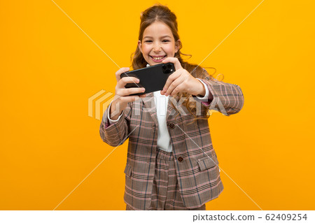 laughing stylish blonde girl in a classic jacket holds a smartphone and plays a game on a yellow 62409254